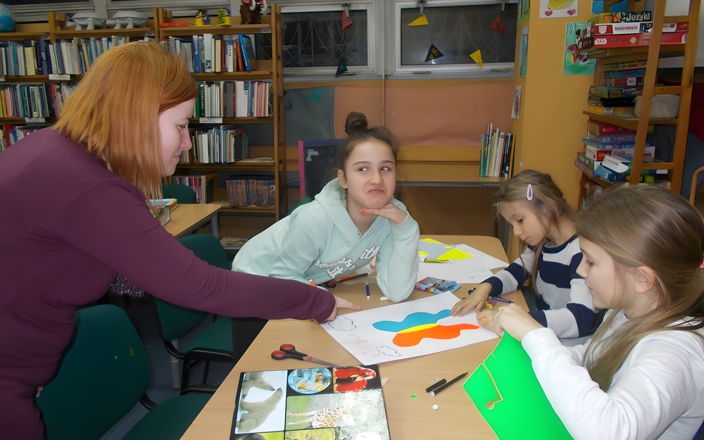 Dzieci siedzą przy stole i pracują w grupie, a bibliotekarka wspiera ich pracę
