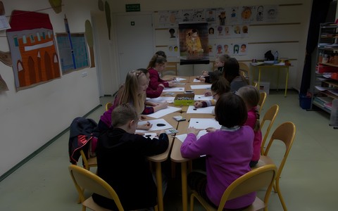 Zdjęcie zrobione jest trochę z góry i obejmuje grupę dzieci siedzącą przy stolikach ustawionych razem wzdłuż sali. Każdy ma do dyspozycji kolorowe kredki, pastele olejne i biały papier.