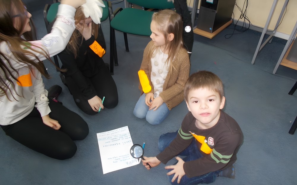 Dzieci siedzą na dywanie w grupie, trzymając lupę i rękawiczkę