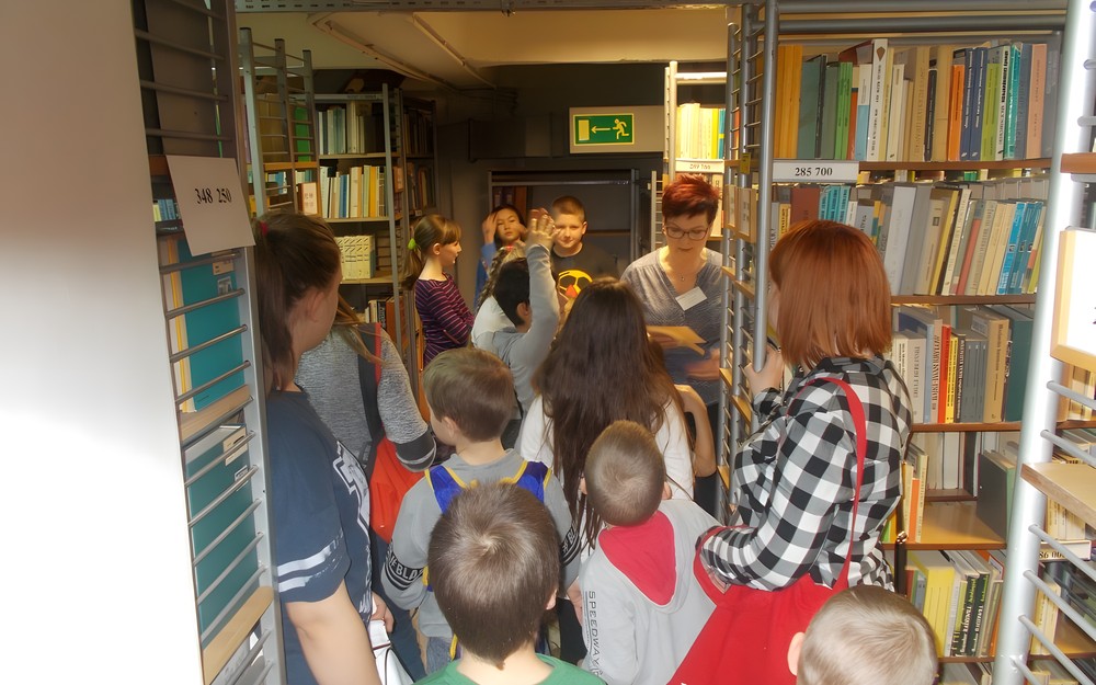 Dzieci z bibliotekarką stoją w magazynie przy regałach