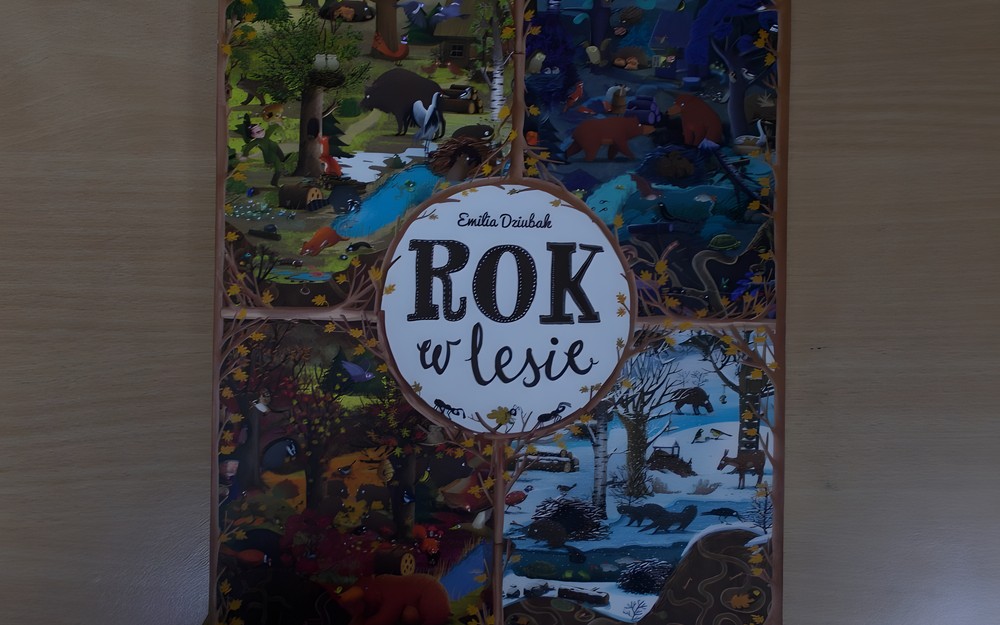 Okładka książki „Rok w lesie” Emilii Dziubak. Tytuł ksiązki i autorka umieszczone są w kółku, pośrodku okładki, tło kółka jest białe, napis ciemnobrązowy. Okładka posiada cztery prostokątne ilustracje z różnymi zwierzętami mieszkającymi w lasach.