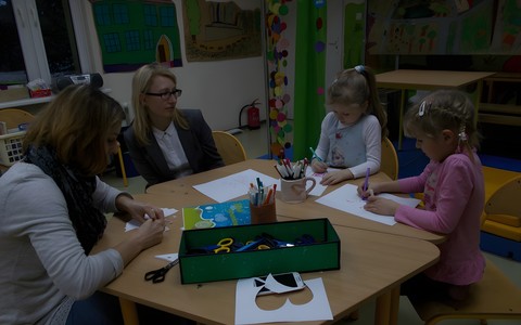 Sala Muminków. Przy stoliku siedzą dwie dziewczynki i dwie panie. Dziewczynki po prawej stronie zajęte są malowaniem motyli. Na stole ustawione są dwa gliniane kubki z flamastrami. Przed nimi podłużny karton z nożyczkami.