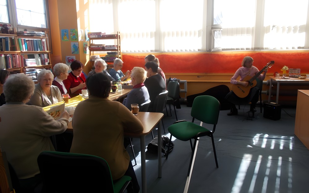 Panie z bibliotekarką siedzą przy stole i rozmawiają, a pani z boku gra na gitarze