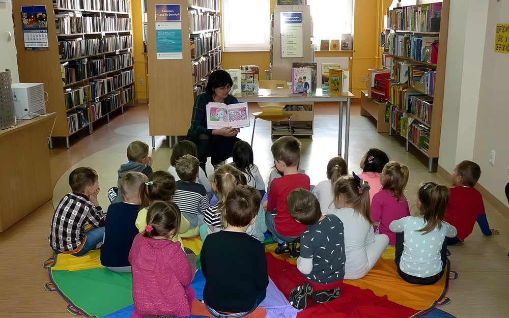 Dzieci siedzą na chuście animacyjnej, a bibliotekarka prezentuje książki o zabawkach