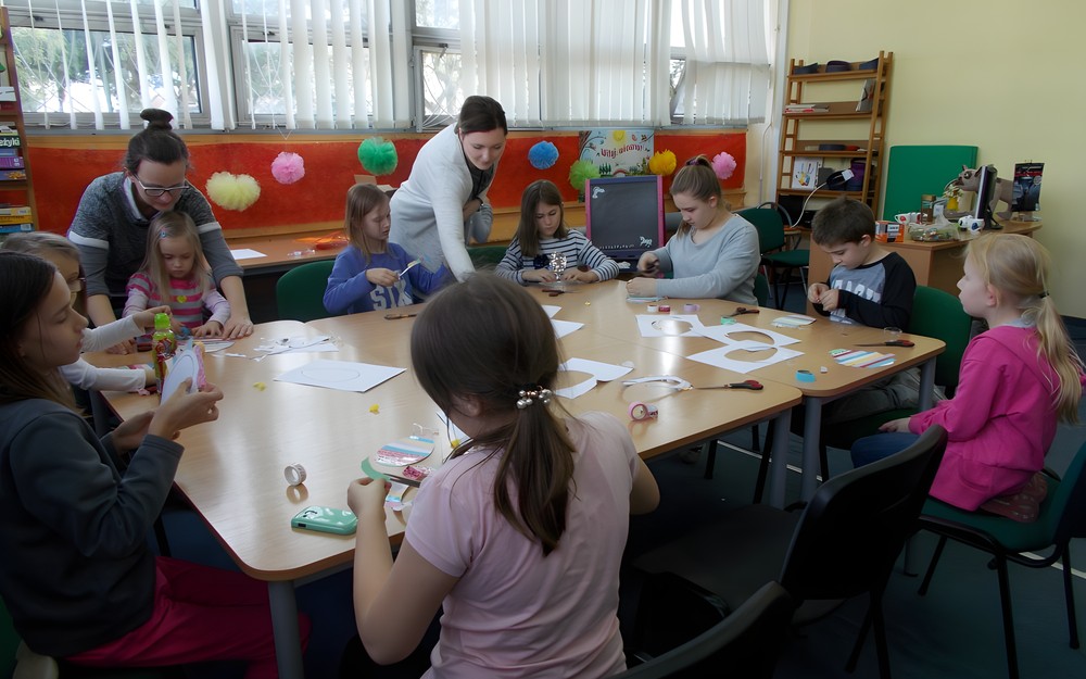 Dzieci wykonują pracę plastyczną przy stole, a bibliotekarka wraz z rodzicem wspomaga ich pracę