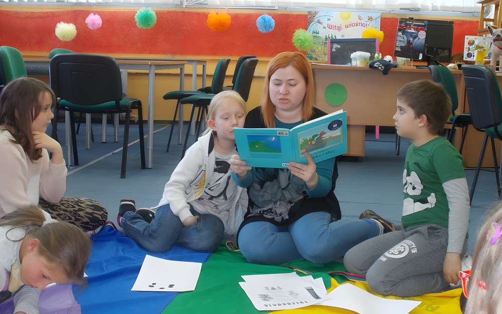 Bibliotekarka siedzi z dziećmi na dywanie i czyta im książkę