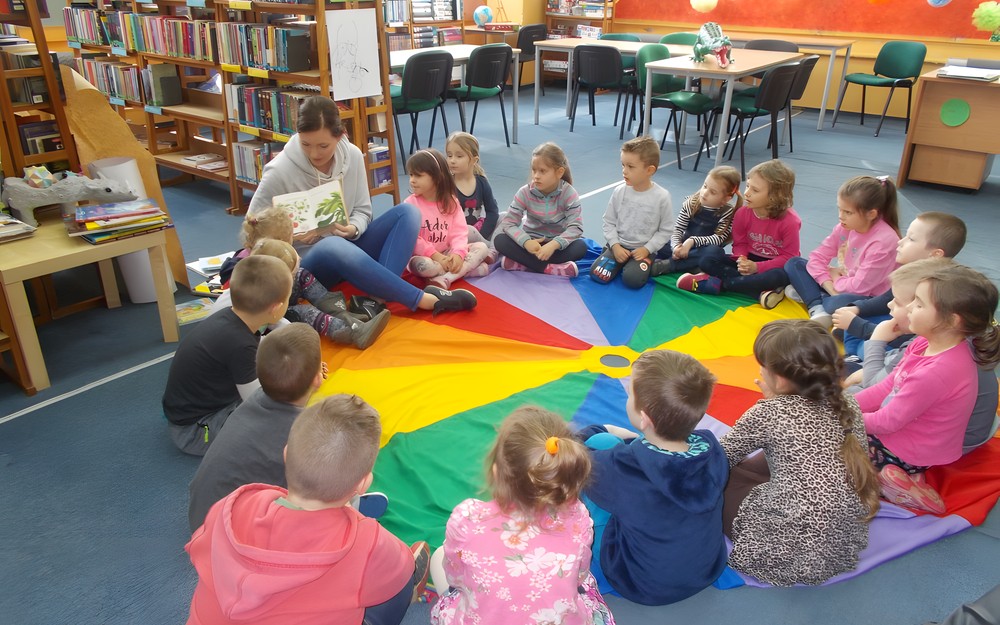 Bibliotekarka siedzi z dziećmi na chuście i czyta im książkę