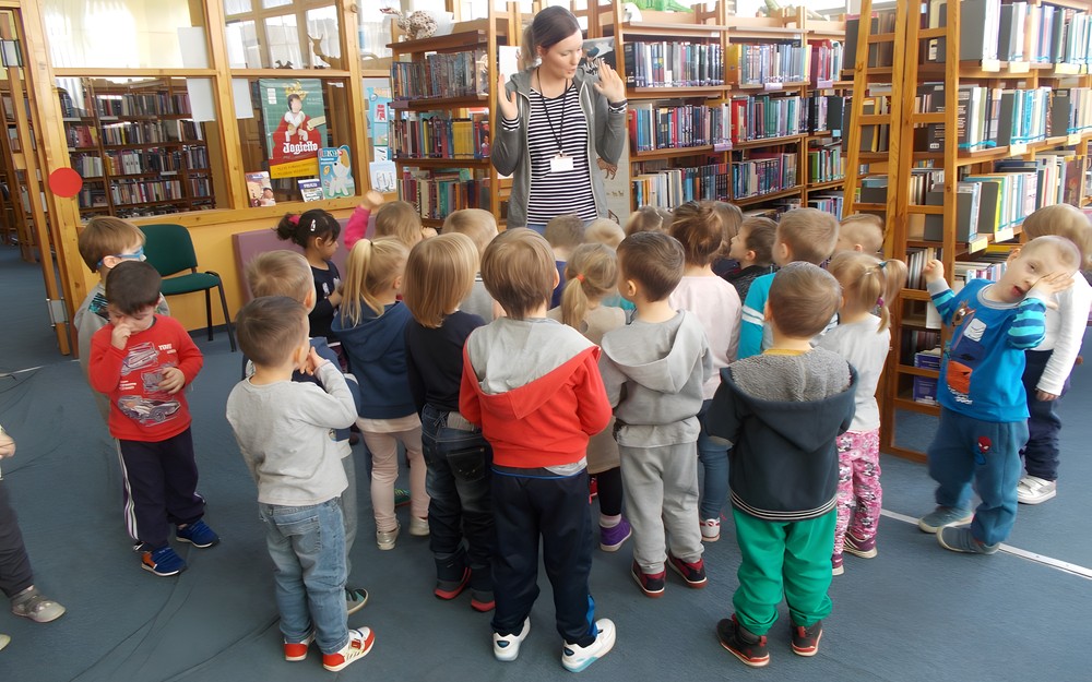 Dzieci stoją na przeciwko bibliotekarki i słuchają opowieści