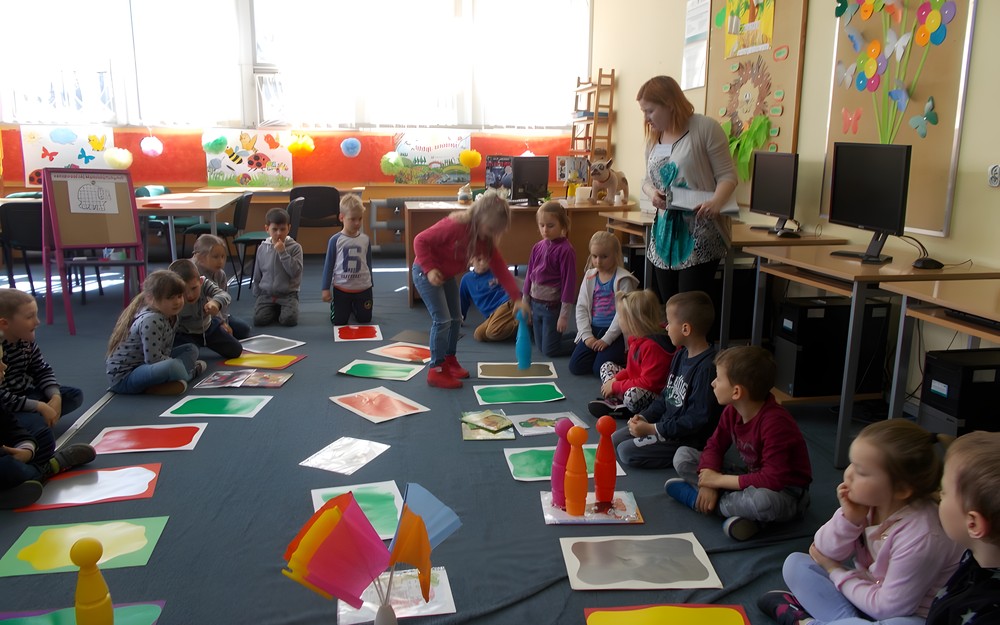 Dzieci siedzą w kole na około gry planszowej, a dziewczynka porusza pionkiem po planszy.