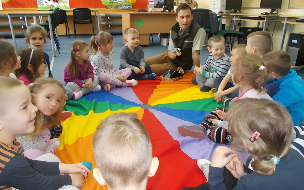 Leśniczy siedzi z dziećmi na dywanie i trzyma w ręku pluszową maskotkę ptaka