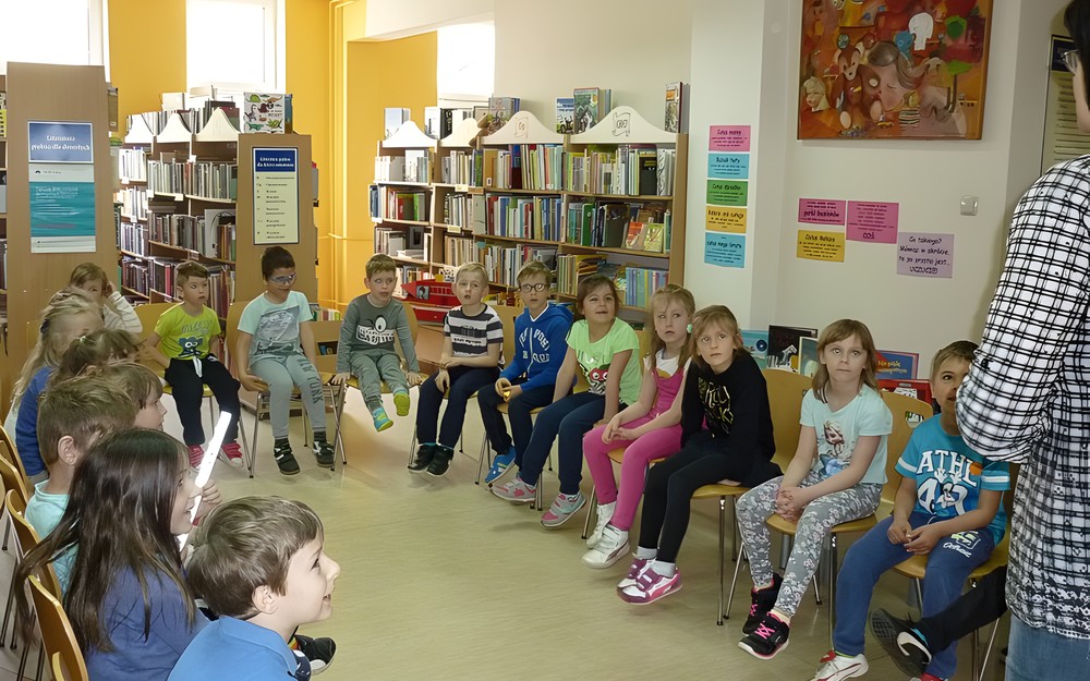 Dzieci siedzą na krzesełkach w kręgu i w skupieniu słuchają bibliotekarki