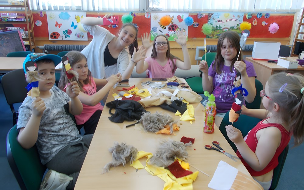 Dzieci  siedzą przy stoliku i pozują do zdjęcia z pacynkami