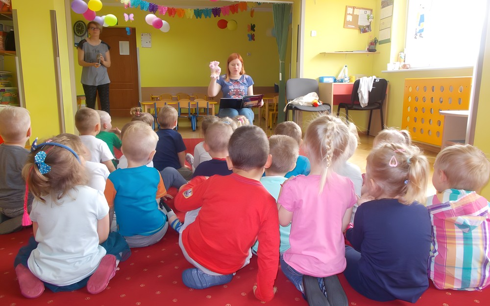 Dzieci siedzą przed bibliotekarką, która czyta książkę i trzyma pacynkę świnkę