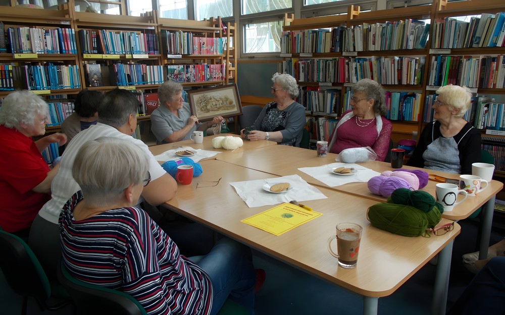 Panie siedzą przy stołach, na których jest dużo włóczek, a jedna z pań pokazuje wyszyty własnoręcznie obraz