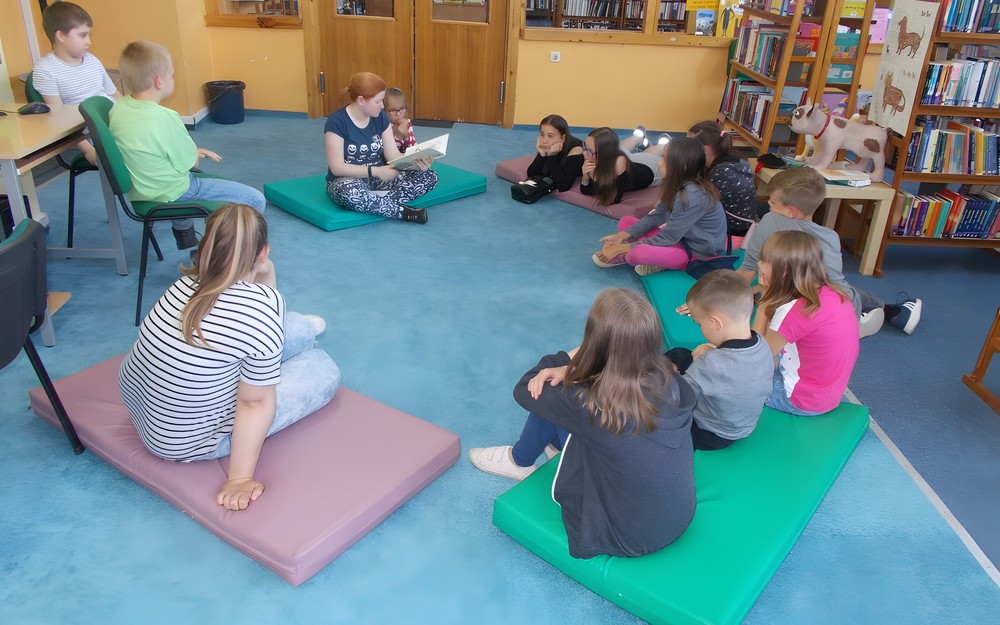 Dzieci siedzą na materacach podczas gdy bibliotekarka wyjaśnia dzieciom zasady zabawy