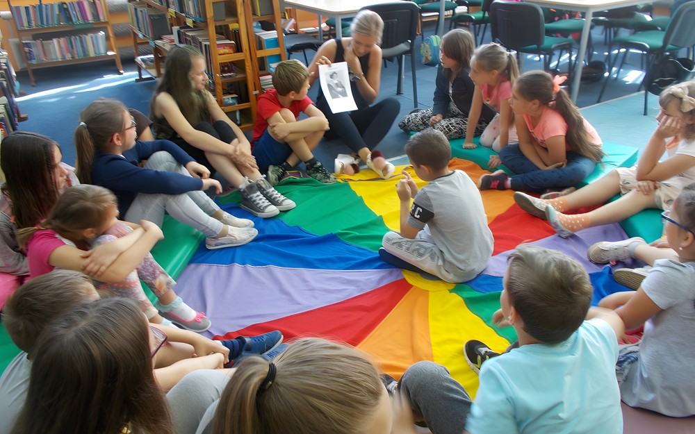 Dzieci siedzą na chuście animacyjnej, a bibliotekarka pokazuje zdjęcie Krzysztofa Kolumba