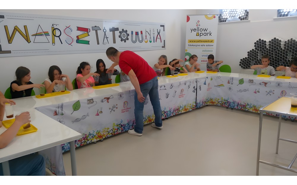 Dzieci siedzą przy stołach słuchając prowadzącego warsztaty chemiczne