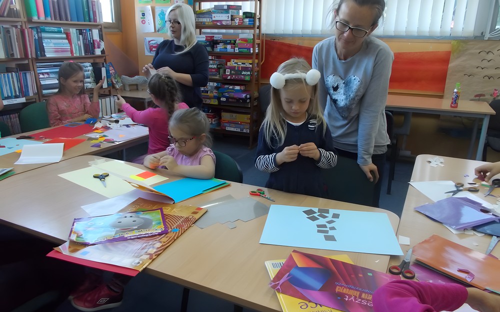 Dzieci siedzą przy stole i tworzą obrazy, a rodzice wspierają ich prace