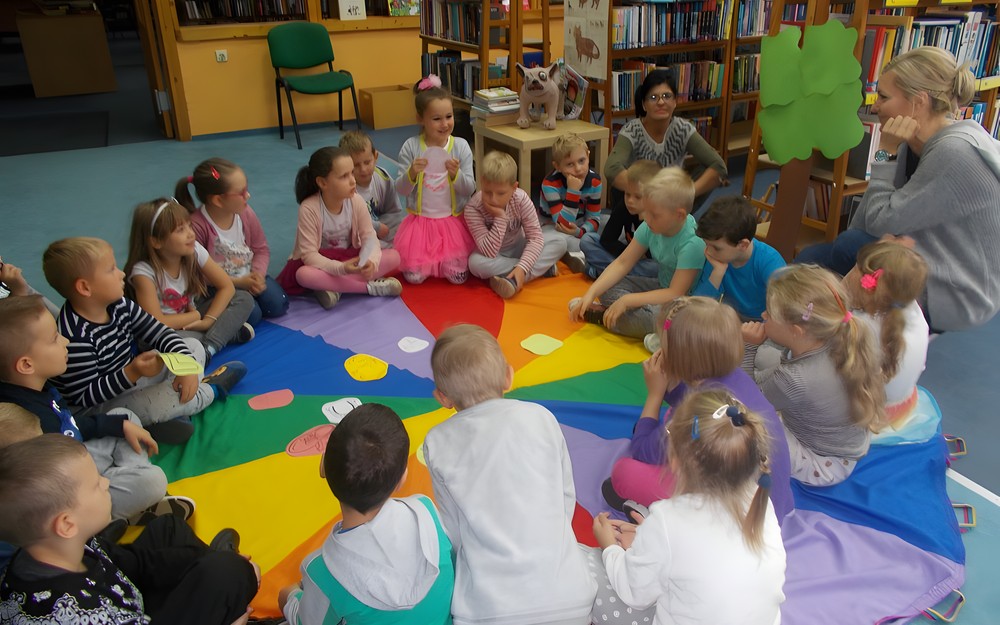 Dzieci siedzą w kole na chuście animacyjnej, na środu leżą symbole szczęścia