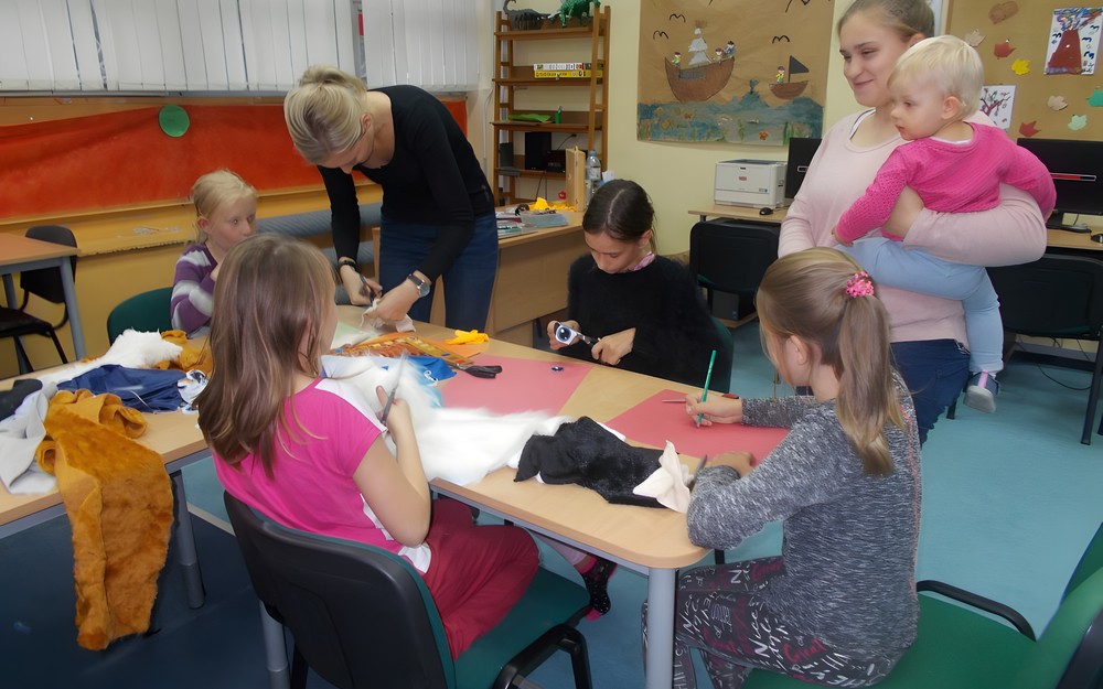 Dzieci siedzą przy stole i wykonują prace plastyczne, a bibliotekarka wspiera ich pracę