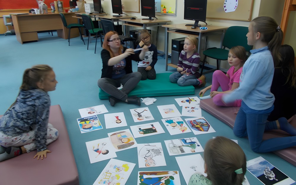 Dzieci siedzą na materacach i patrzą na bibliotekarkę trzymającą karteczkę z nazwą zawodu po angielsku