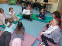 Dzieci siedzą naokoło bibliotekarki, która czyta im opowieść