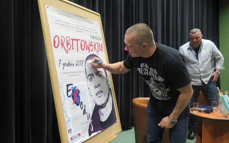 Łukasz Orbitowski składa pamiątkowy autograf na plakacie promującym spotkanie