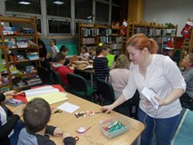 Dzieci siedzą przy stołach i wykonują pracę plastyczną a bibliotekarka koordynuje ich pracę