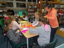 Dzieci siedzą przy stole i wycinają elementy świąteczneych symboli a rodzice wspierają ich pracę