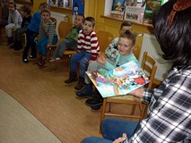 Dzieci siedzą na krzesełkach, słuchają czytanej przez bibliotekarkę książki o Mikołaju