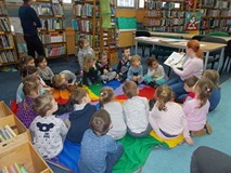 Dzieci siedzą na chuście animacyjnej i słuchają opowieści czytanej przez bibliotekarkę