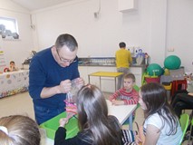 Prowadzący trzyma worek wypełniony wodą z wbitymi kredkami, a dziewczynka dokłada kolejną