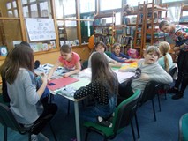 Dzieci siedzą przy stole i wykonują pracę plastyczną, a bibliotekarka wspiera ich w działaniu