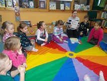 Dzieci siedzą na chuście i patrzą w stronę bibliotekarki, która trzyma w ręku książkę