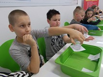 Dzieci siedzą przy stołach i wykonują doświadczenia