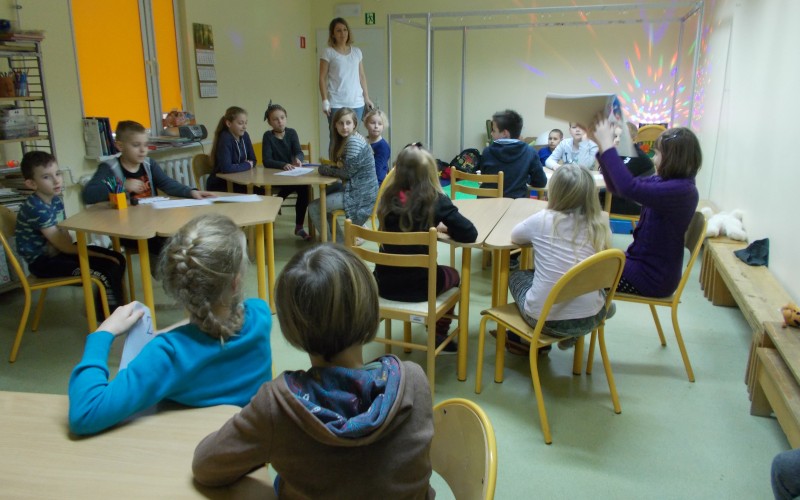 Dzieci siedzą przy stolikach słuchając bibliotekarki, która opowiada im ciekawostki o kosmosie