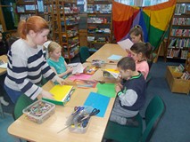 Dzieci siedzą przy stole i wykonują prace plastyczne, a bibliotekarka wspiera ich pracę