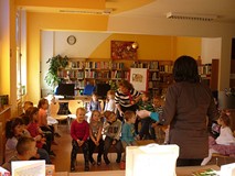 Bibliotekarka wita dzieci siedzące na krzesełkach i ławeczkach