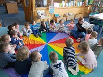 Dzieci siedzą na chuście i słuchają bibliotekarki, która czyta im książkę