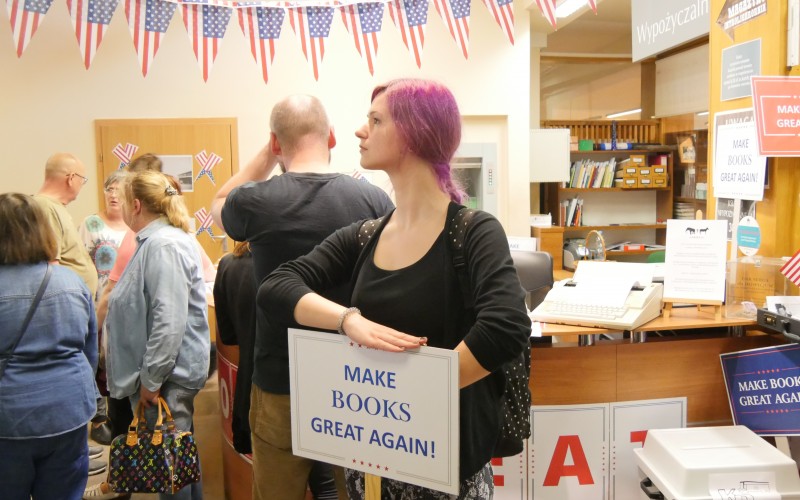 W centrum zdjęcia widoczna młoda kobieta w rózowych włosach z tabliczką Make books great again!