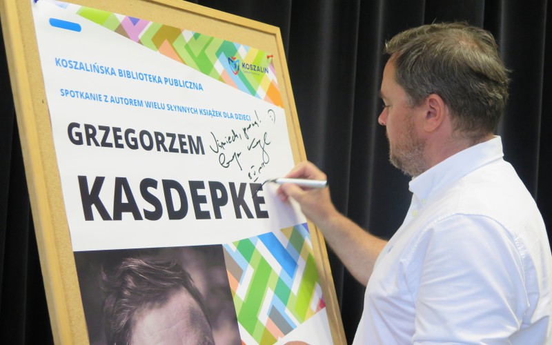 Grzegorz Kasdepke składa pamiątkowy autograf na plakacie reklamującym spotkanie