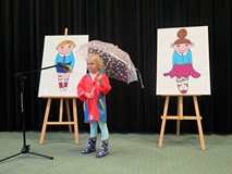 Dziewczynka recytuje wiersz. Ubrana jest w pelerynę i ma w reku parasol.