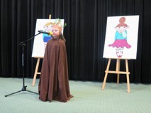 Cziewczynka w brązowej pelerynie, z koroną z drzew na głowie recytuje wiersz