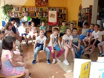 Dzieci siedzą na ławeczce i krzesełkach. Słuchają czytanych przez bibliotekarkę książeczek