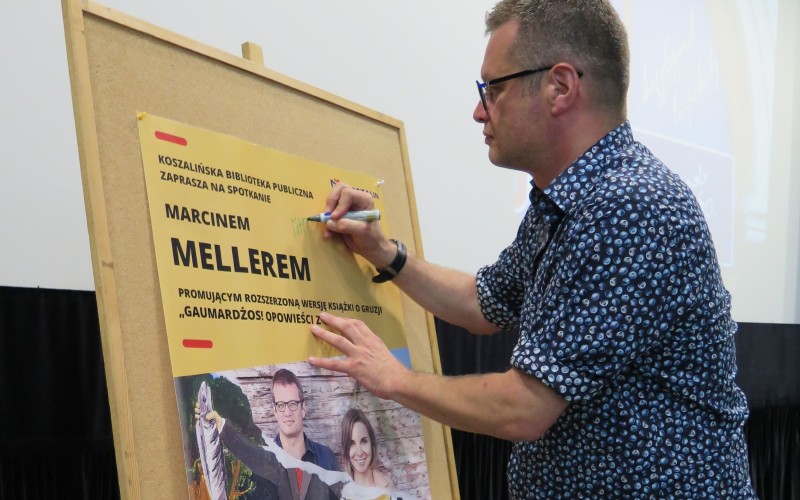 Marcin Meller składa pamiątkowy autograf na plakacie promującym spotkanie
