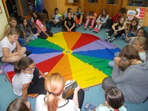 Dzieci siedzą na chuście animacyjnej, a bibliotekarka pokazuje im ilustracje z książki