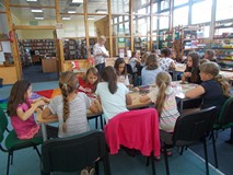 Dzieci siedzą przy stole podczas gsy prowadzaca wyjaśnia zasady pracy plastycznej