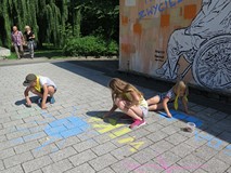 Dwie dziewczynki i jeden chłopiec malują kredą przed budynkiem głównym Koszalińskiej Biblioteki Publ