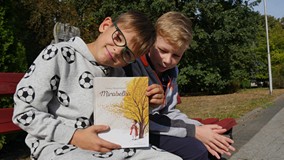 Dwaj chłopcy siedzą na ławce. Jeden z nich prezentuje książkę Cezarego Harasimowicza  „ Mirabelka – 