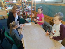 Dzieci siedzą przy stole i wykonują prace plastyczną, a bibliotekarz wpsomaga ich pracę
